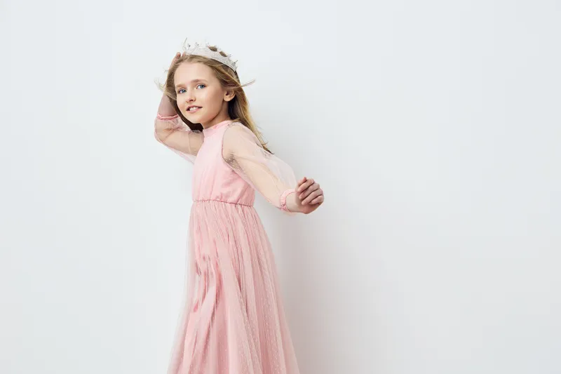 Delicate young girl in a light pink dress with a floral crown, posing playfully against a soft white background, epitomizing joy and innocence during a spring celebration