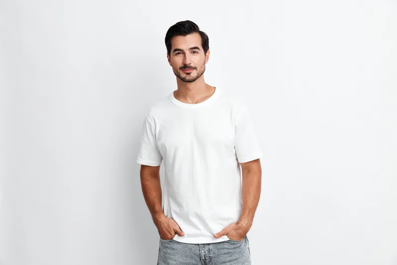 Smiling young man wearing a white t-shirt poses confidently with hands in pockets against a plain solid background. Casual style and relaxed vibe concept.