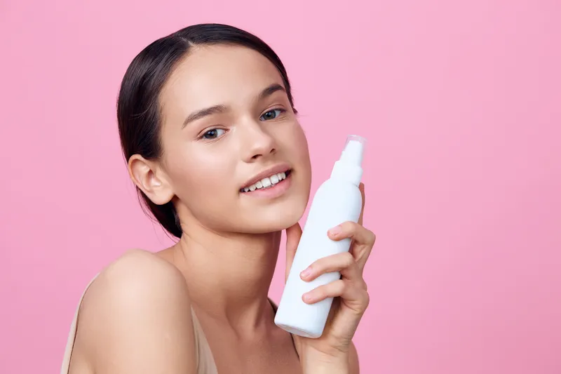 Young woman holding a skincare product with a cheerful expression, against a pastel pink background, promoting healthy skincare routines and self care