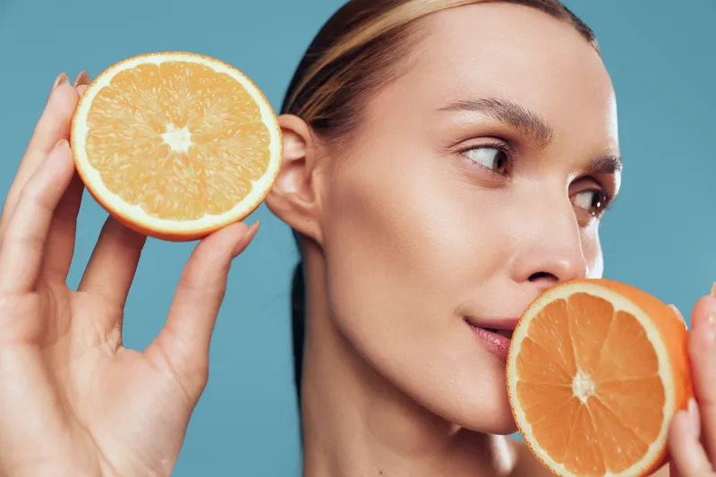 A young woman with clear skin holds two halves of a fresh orange near her face against a blue background. Concept for healthy lifestyle, natural beauty, and skincare with citrus fruit.