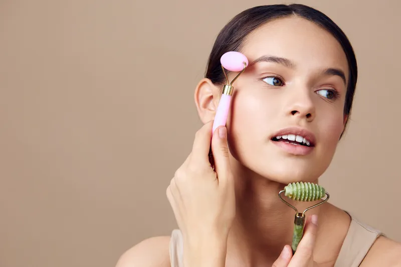 Young woman using jade roller and facial massager for skincare routine, relaxed expression against a neutral background, healthy beauty concept