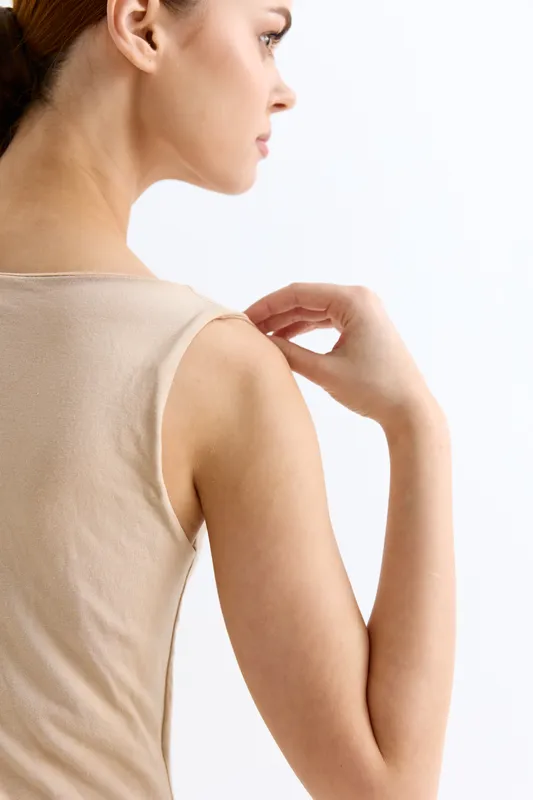 young woman in beige sleeveless top touching her shoulder with a gentle expression against a clean white background, highlighting skin care and casual style