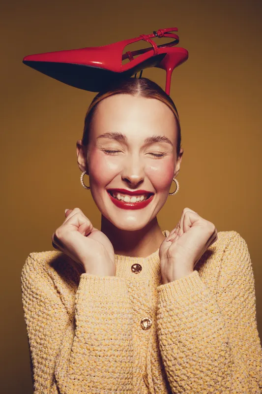 Happy woman with red high heel shoe on head smiles with closed eyes and clenched fists. She wears yellow knitted sweater and gold hoop earrings on brown background. Concept of joy and fashion.