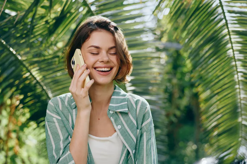 cheerful woman outdoors talks on a phone, smiling softly in a sunny tropical garden setting with lush palm leaves