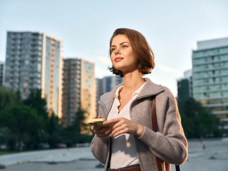 A confident professional woman stands outdoors in an urban cityscape, checking a smartphone. She wears a light jacket and blouse, smiling softly as daylight highlights her features, projecting a