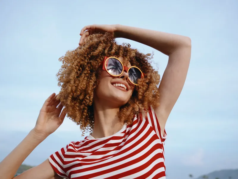 Joyful young woman with curly hair wearing a red and white striped shirt and round sunglasses, smiling brightly as she enjoys sunny outdoor moments.