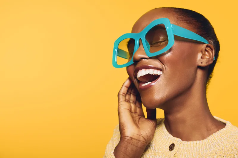 Smiling black woman with dark skin wearing large turquoise sunglasses and beige sweater poses against yellow background. Concept of fashion, joy, summer style, and confident beauty.