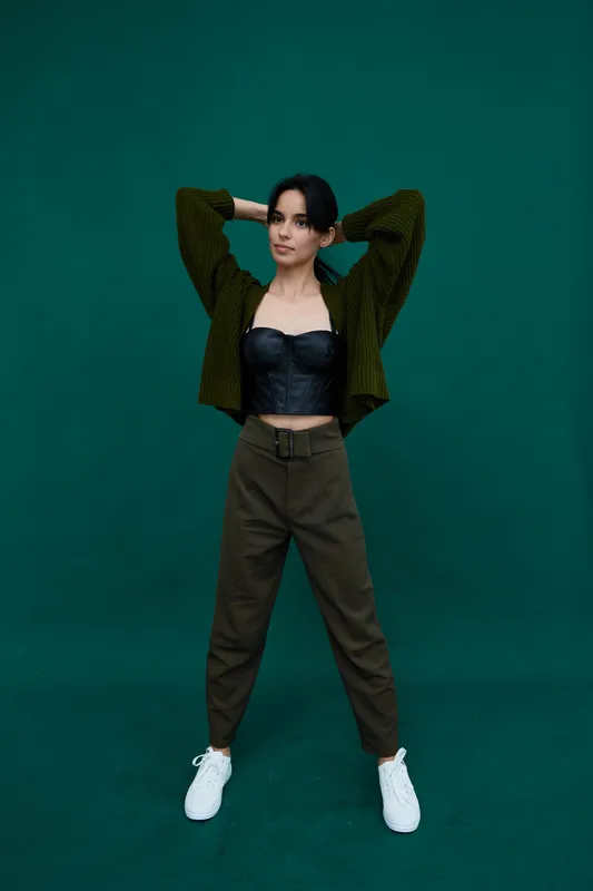 Young woman posing confidently in casual green cardigan and dark pants against a solid green background, wearing white sneakers and black strapless top.