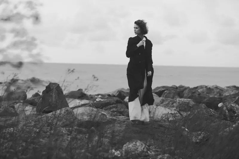 Woman on a rocky shoreline in a long coat, gazing toward the sea, moody black and white scene with wind swept coast, fashion and solitude in nature