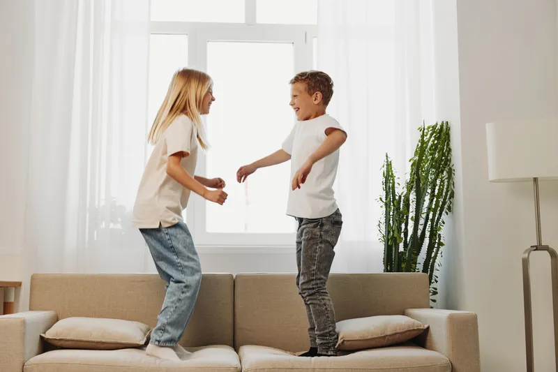 Two happy children jumping on beige sofa in bright living room with large window and green plant. Concept of childhood joy, playful siblings, and home fun activity indoors.