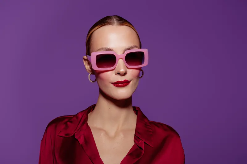 Stylish woman with pink sunglasses and red lipstick poses confidently against purple background. She wears a red blouse and gold hoop earrings. Concept of fashion, beauty, and modern style.