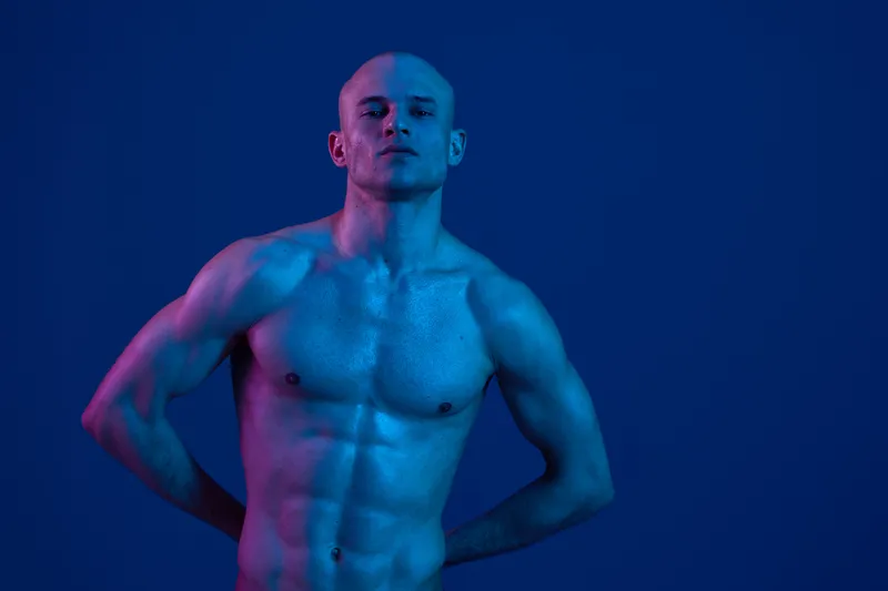 Muscular man with glistening skin posing confidently in a captivating blue light, showcasing strength and elegance in a stunning black and white image.