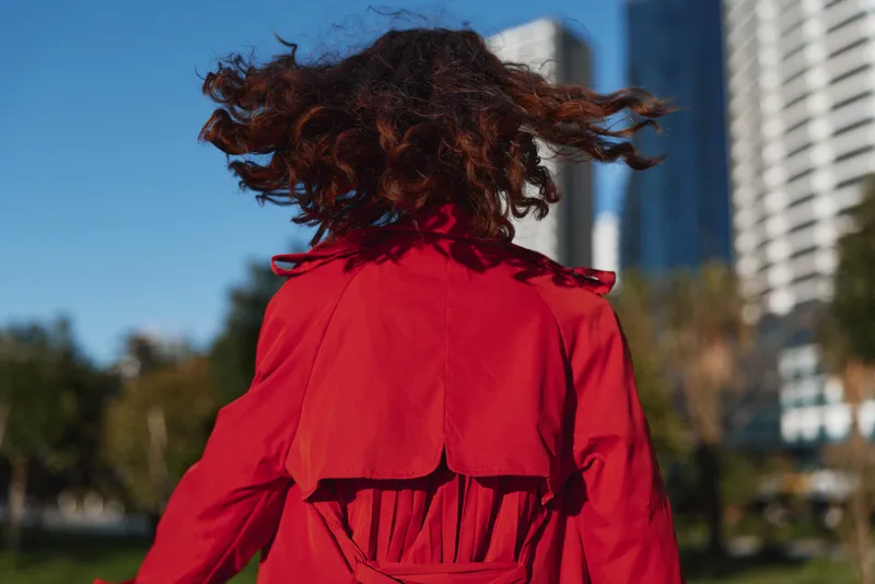 A young woman with curly red hair wears a bright red coat while spinning outdoors in an urban park on a clear sunny day.