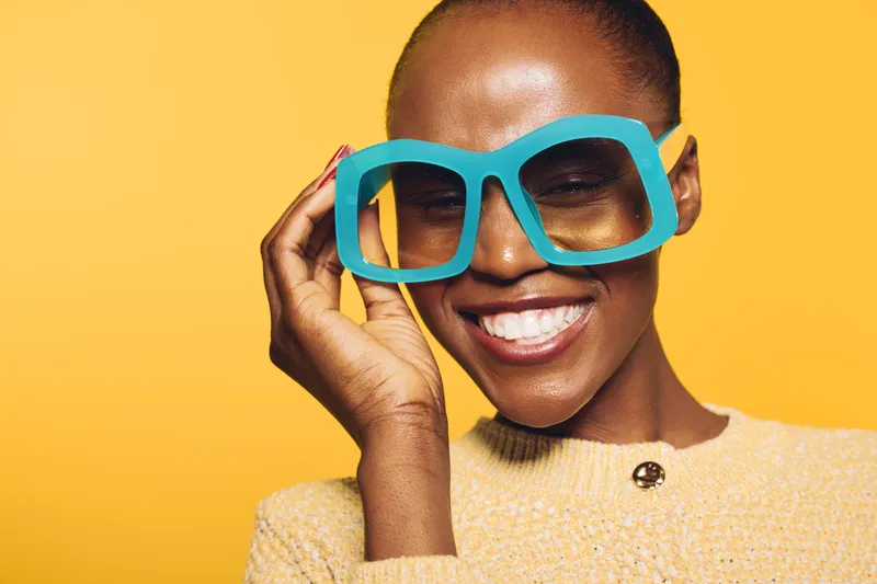 Smiling black woman with dark skin wearing oversized blue glasses and yellow sweater. Close-up on yellow background. Concept of fashion, happiness, and joyful emotions in studio lighting.