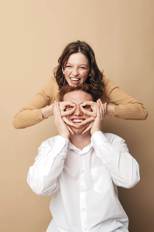 Happy young couple having fun together, woman playfully covering man’s eyes with her hands. Casual clothing, beige background, joyful mood, friendship and love concept in studio lighting.