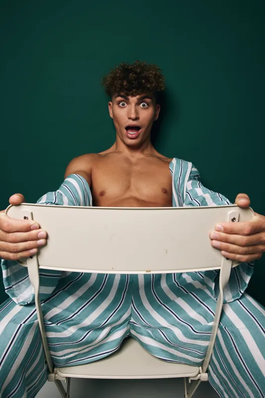 Young man with curly hair striped pajamas holding a beige chair excited and surprised sitting against green backdrop. His expression is wide-eyed emotion and shocking sensation indoors.