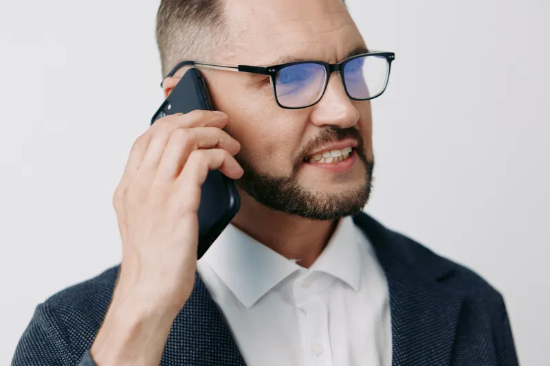 A professional man on a solid colored background, speaking on a mobile phone with a focused, confident expression, conveying business communication and urgency.