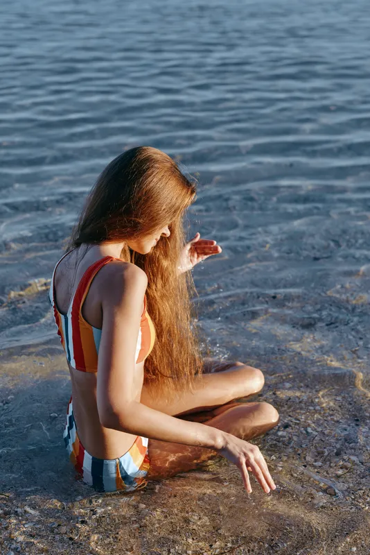 Young woman sitting on beach sand near ocean waves, striped swimsuit, long hair catching sunlight, relaxed summer vacation mood, natural outdoor light, close view