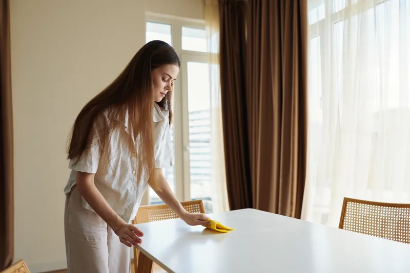Woman cleaning table at home while talking on phone, wiping surface with yellow cloth in sunlit dining room. Domestic chores, tidy interior and everyday household routine.