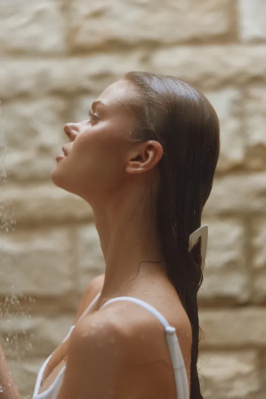 Profile of a young woman under shower water with wet hair, peaceful expression, and soft natural light against textured stone background in spa setting.