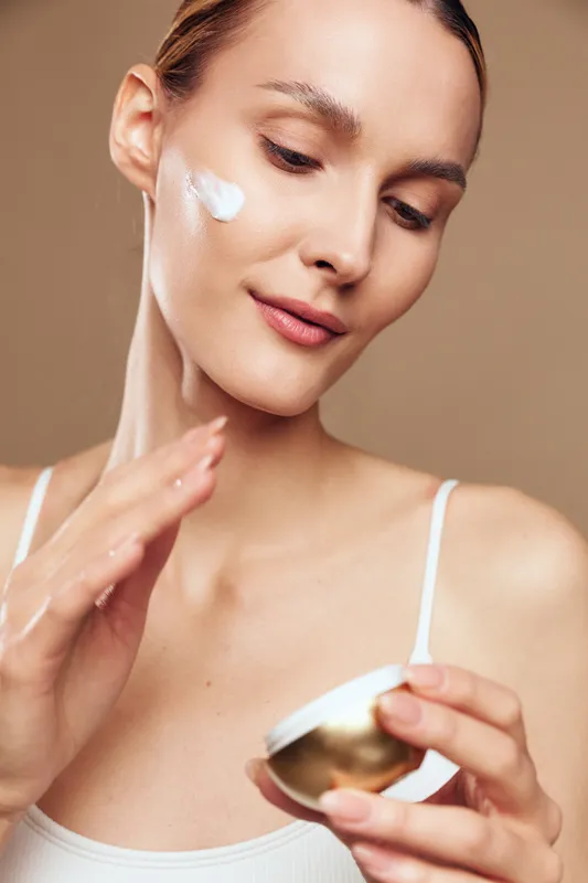 A young woman applies face cream on her cheek with a gentle smile, wearing a white tank top against a beige background. Concept for skincare, beauty routine, and self-care.