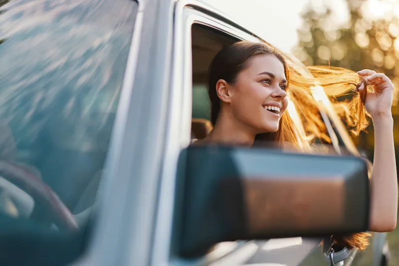 Young woman enjoying a road trip, smiling and playing with her hair, sunlight shining through a car window, capturing the essence of freedom and adventure.