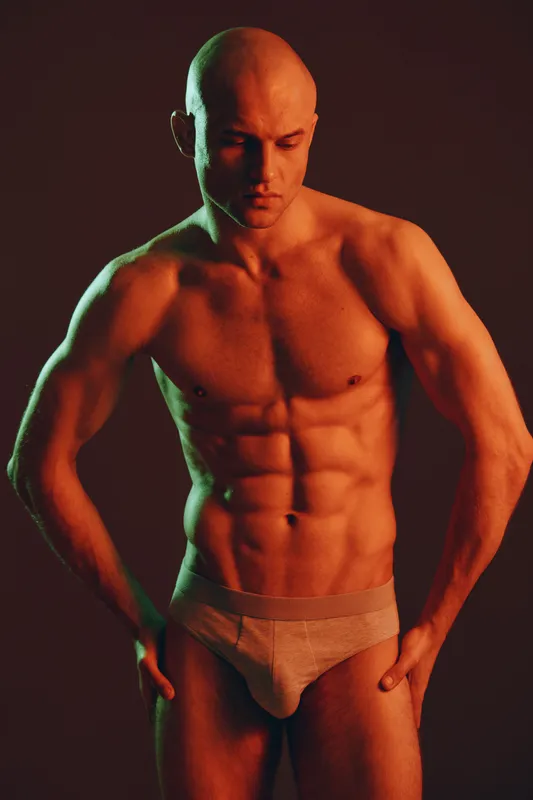 Muscular man poses in minimalistic style, showcasing his physique under dramatic lighting in a black and white aesthetic, emphasizing strength and confidence.