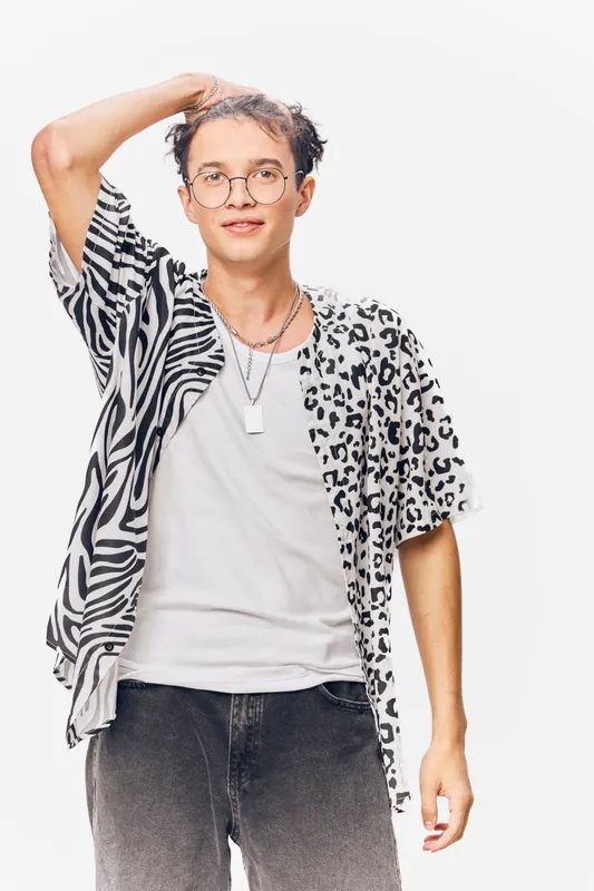 Young man wearing mixed animal print shirt and glasses, posing confidently with a hand in hair against white background for modern fashion style.