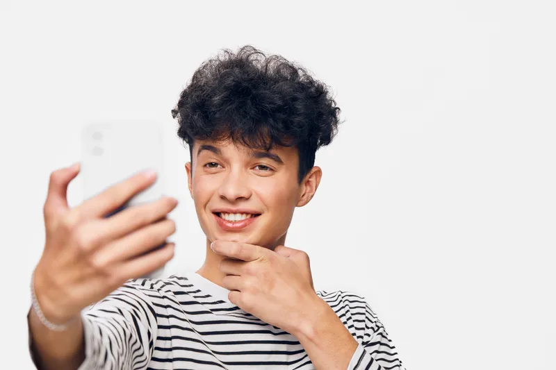 Young man smiling confidently taking a selfie with smartphone, curly hair and striped shirt on a bright white background, casual and modern lifestyle image.