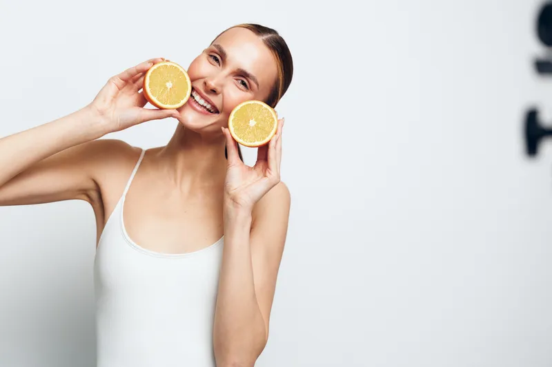 Smiling woman holding two halves of fresh orange near face on white background. Concept of healthy lifestyle, natural beauty, and vitamin C. Bright studio lighting creates cheerful mood.