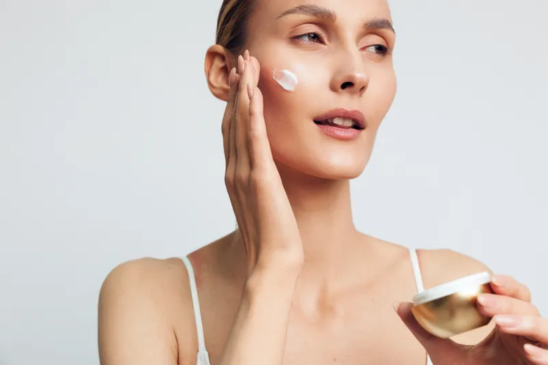 Close up of a woman applying moisturizer to her cheek, showcasing a skincare routine with a gentle cream on the skin, soft lighting, natural glow, isolated on a light neutral background