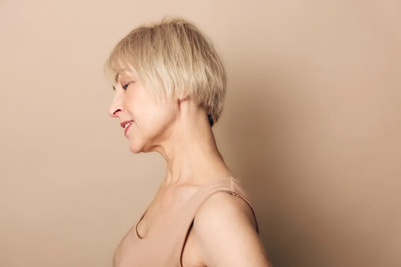 Profile of a mature woman with short blonde hair wearing a beige top against a plain background. Concept of aging, natural beauty, confidence, and serene expression.