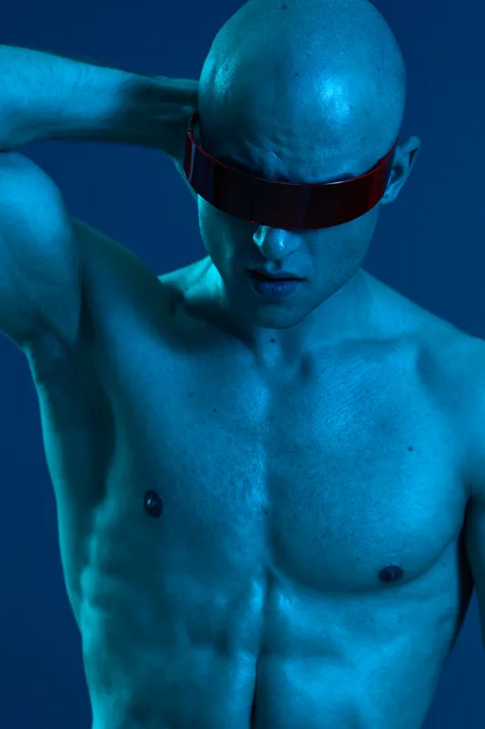 Athletic man with a futuristic look, wearing stylish sunglasses and showcasing a strong physique against a vibrant blue background. Unique and captivating imagery.