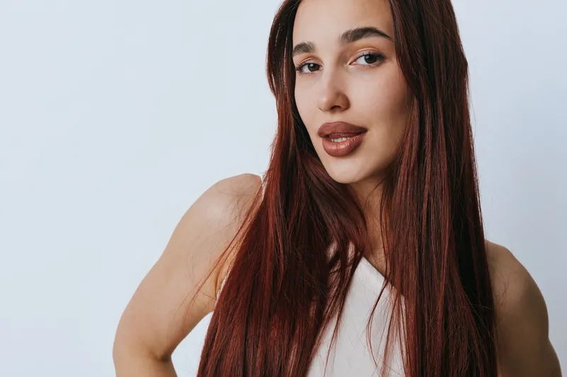 Portrait of young woman with long straight hair and natural makeup posing against plain background. Female model with confident expression wearing sleeveless top in studio light.