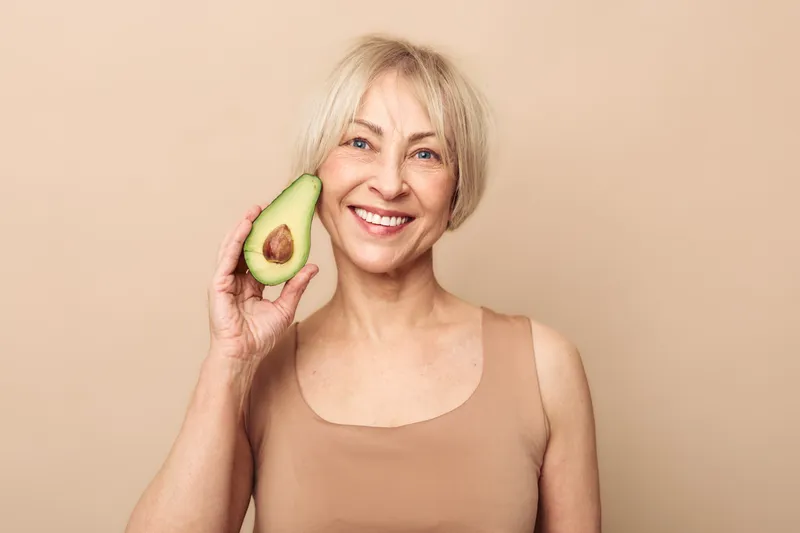 Smiling mature woman with short blonde hair holds half avocado near face, wearing beige tank top on neutral background. Concept of healthy lifestyle, nutrition, wellness, and natural beauty.