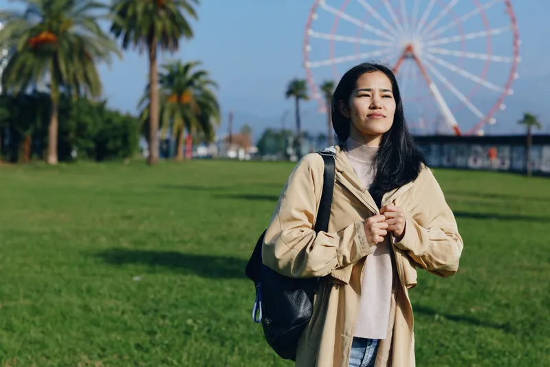 Woman in beige coat standing in a sunny park with a backpack, smiling softly, ferris wheel visible in the distance, casual outdoor moment perfect for travel, leisure, and everyday photography