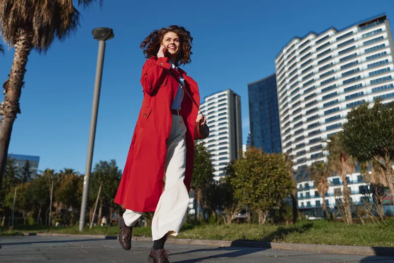 A stylish woman in a bright red coat walks along a sunny city park with modern high rises in the background, exuding confident urban energy and casual elegance.