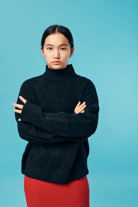 Young woman with serious expression wearing black turtleneck sweater and red skirt standing with arms crossed against blue background in studio, indoor portrait lighting and style.
