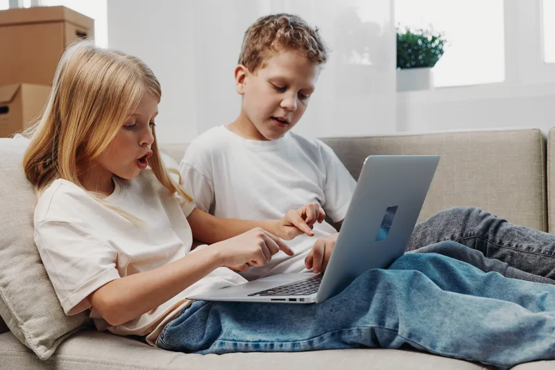 Two children sitting on a couch using a laptop together. The boy and girl are focused on the screen in a bright living room. Concept of learning, technology, and teamwork.
