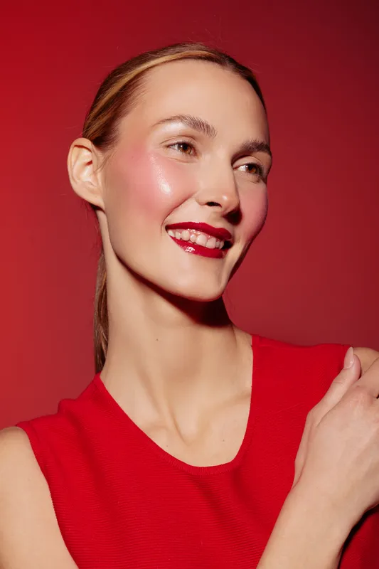 Smiling woman with shiny red lipstick and glowing skin wears red sleeveless top against red background. Concept of beauty, makeup, skincare, fashion, and confident female portrait.