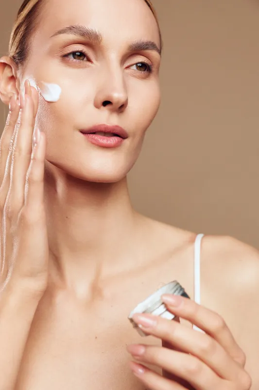 Young woman applies cream to her cheek in a calm skincare moment, close up portrait with a soft beige background and gentle lighting, conveying focus, self care and natural beauty