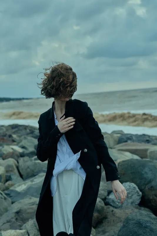 Dramatic windy seaside scene with a figure in a long black coat and white shirt standing on rocks by the sea, conveying movement and moody atmosphere for outdoor fashion