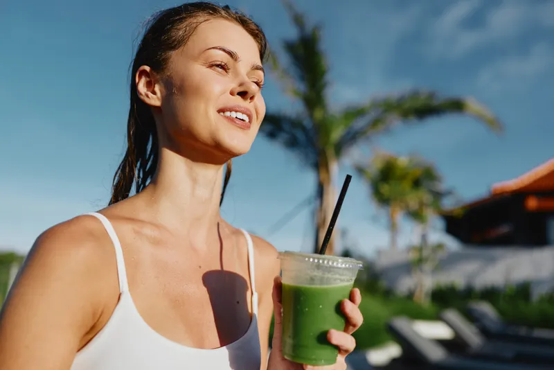 Young woman in white tank top holding green smoothie outdoors, smiling with eyes closed, enjoying sunny day near palm trees. Concept of healthy lifestyle, wellness, and relaxation.