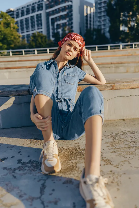 Lifestyle woman in modern boho-western denim street style sitting outdoors, relaxed pose, casual outfit with bandana, film color tones enhancing the urban vibe and warm sunlight.