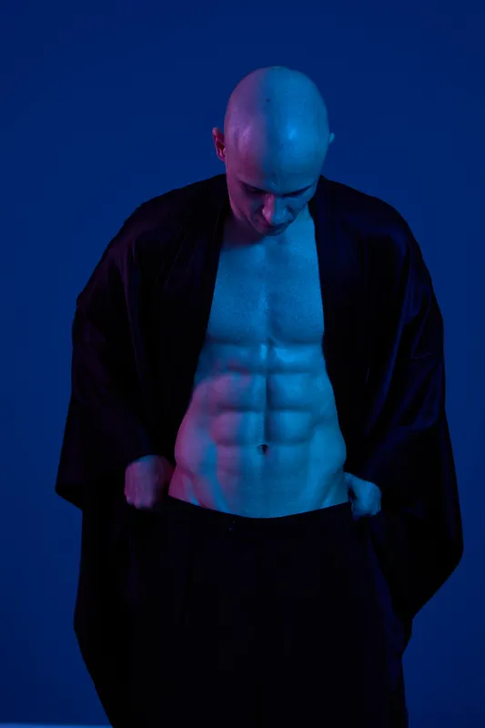 Muscular man showcasing defined abs against a vibrant blue background, wearing a dark robe. Strong presence and bold colors convey fitness and confidence.