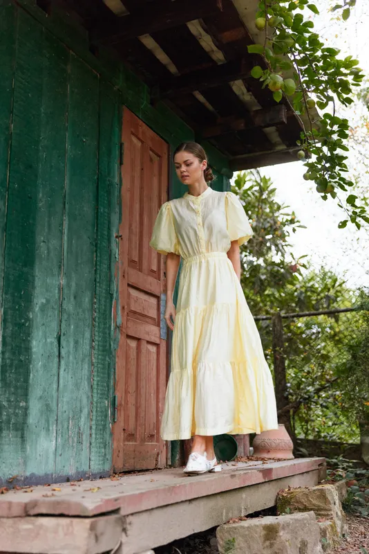 A woman in a yellow dress stands on porch steps beside a green doorway in a garden setting, enjoying natural daylight and a calm, contemplative moment.