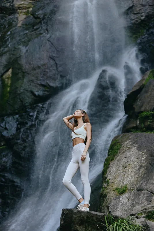 young woman in white activewear stands confidently near a large rocky waterfall in nature, showcasing fitness and outdoor adventure with a peaceful mood