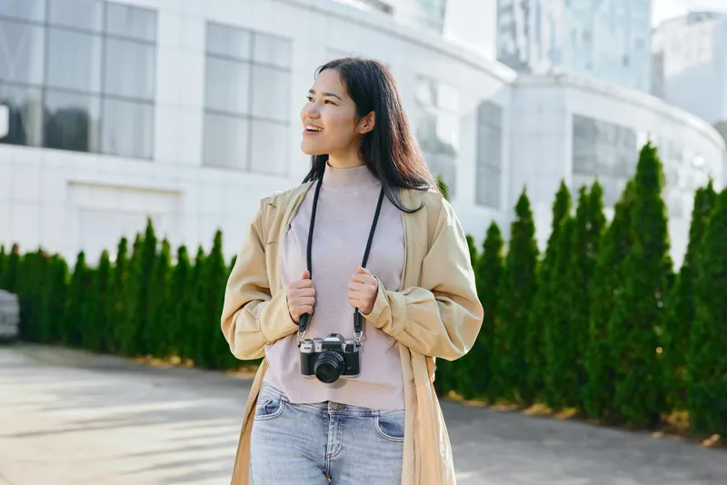 Woman traveler stands outdoor in urban setting with camera hanging, wearing casual jacket and light sweater, modern architecture in background, candid fashion moment, daylight atmosphere and relaxed