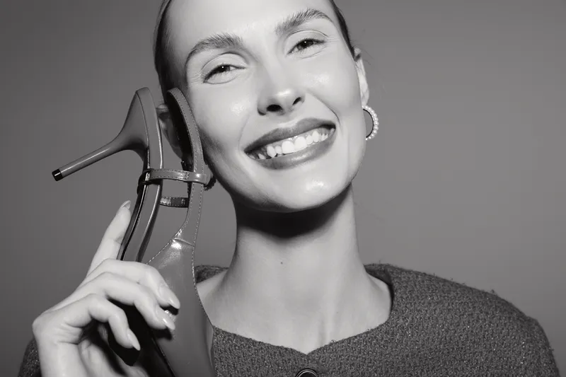 Smiling white woman with fair skin holds high heel shoe near face, wearing earrings and knitted sweater. Studio portrait in black and white. Concept for fashion, happiness, and beauty.