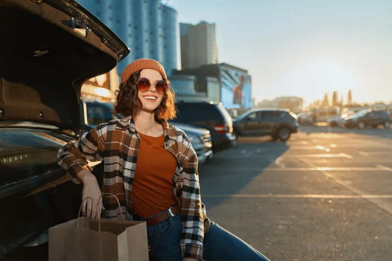 Woman leaning on car trunk with paper bag, shopping and smile during golden hour in urban parking lot, candid lifestyle portrait with authenticity, golden hour glow and emotional storytelling.